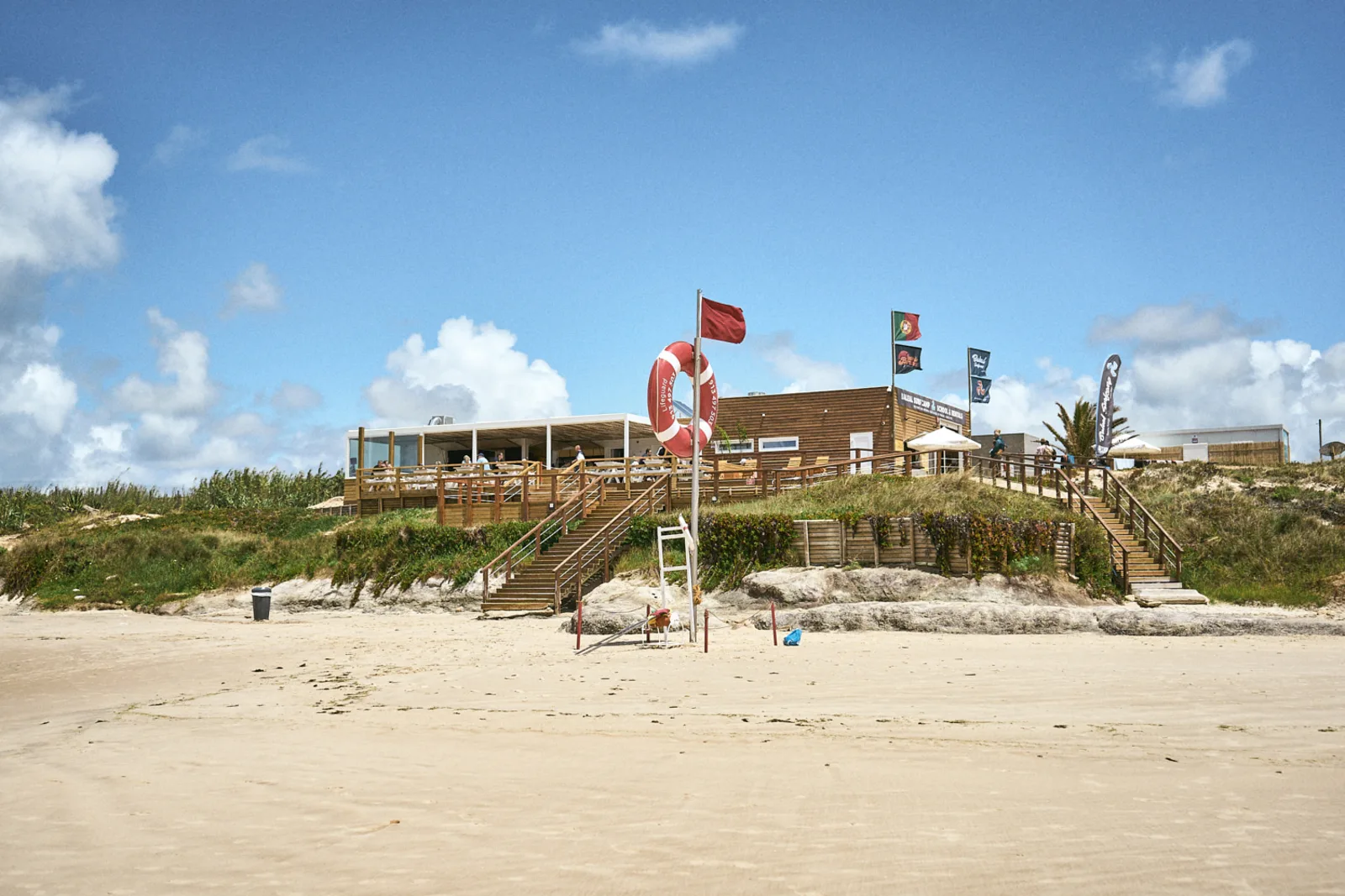 The Baleal Surf Camp surf station and beach bar on the dunes, with a wide sandy beach in front and the Portuguese flag flying